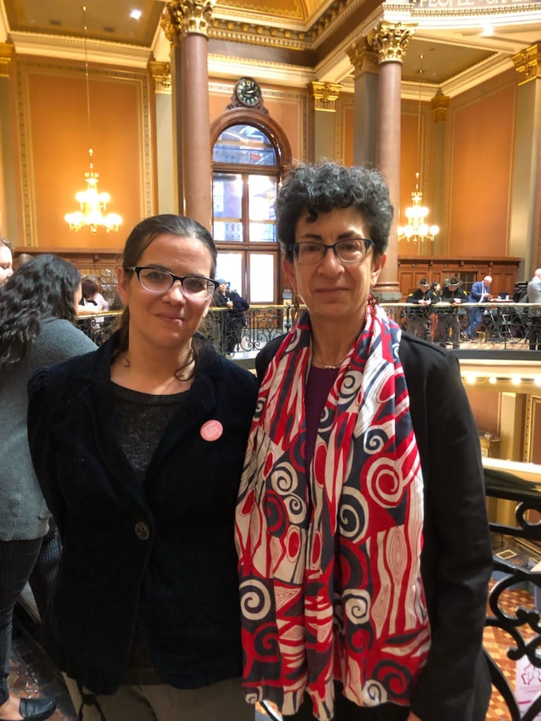 Mandi wearing a black shirt, black buttoned up corduroy coat and glasses with her hair pulled back, standing at the Iowa capitol next to senator Janice Weiner, who is wearing a blue, red, and white scarf and glasses, with short hair.