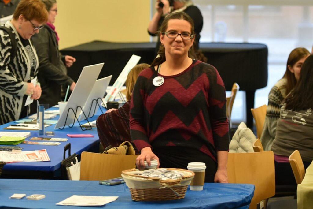 Mandi standing behind a table with a blue cloth, two coffee cups, and a basket of buttons on it at the first CCAN-Con. She is wearing a maroon shirt with black zig-zagged stripes, a CCAN button, and glasses with her hair pulled back.