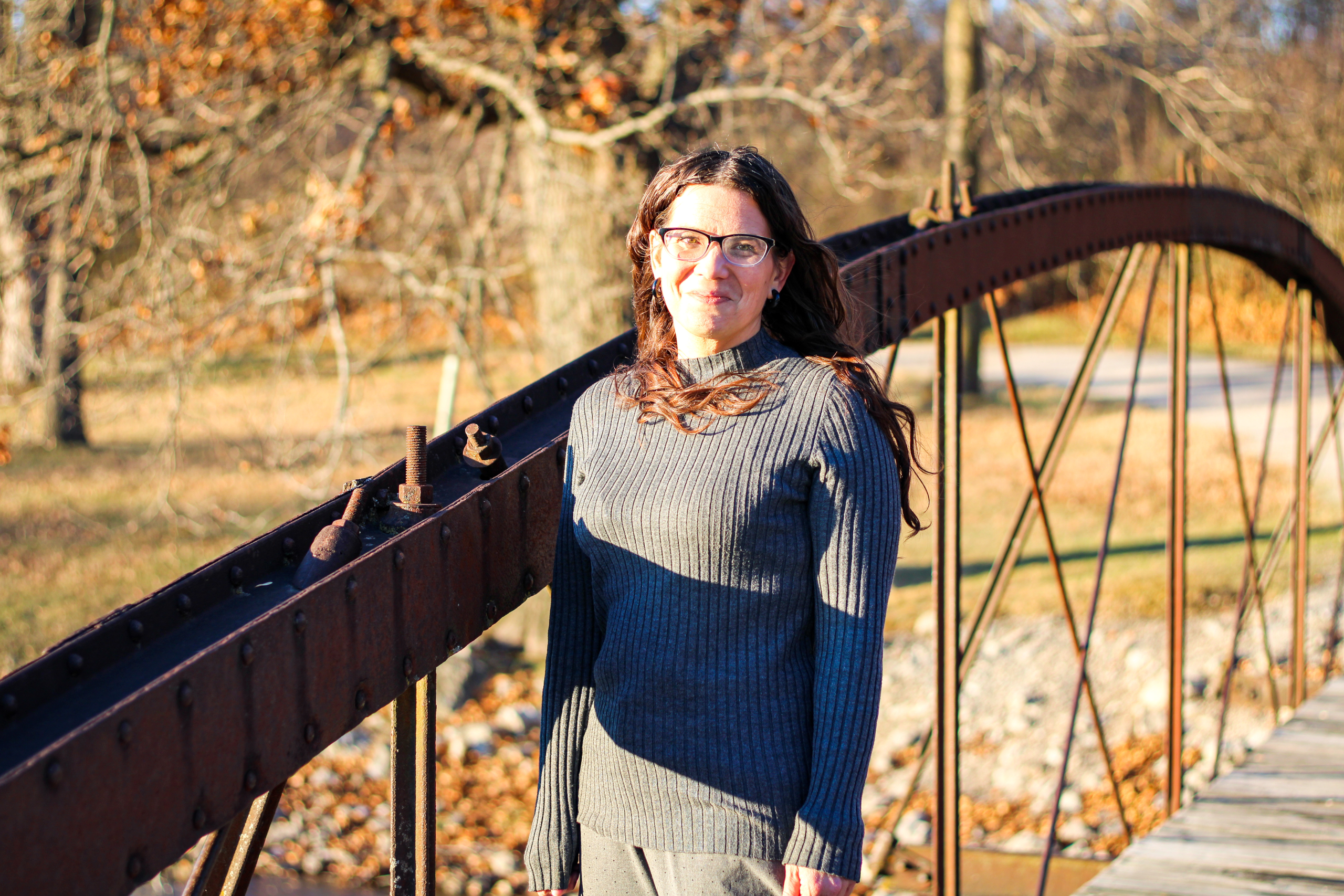 Mandi standing on a bridge. She's wearing a gray sweater and smiling, and her hair is blowing slightly to the right. It's autumn and there are orange leaves in the background.